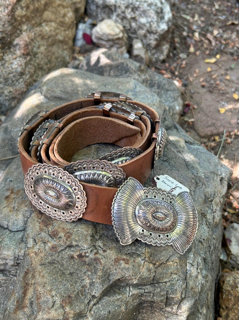 May include: A brown leather belt with ornate silver-colored metal embellishments. The belt features oval and butterfly-shaped buckles with intricate designs. The belt is displayed on a textured rock surface.