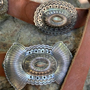 May include: Close-up of a brown leather belt with three silver-colored oval belt buckles. The buckles have intricate engraved designs with a central eye-shaped element and sunburst patterns. The belt is resting on a textured stone surface.