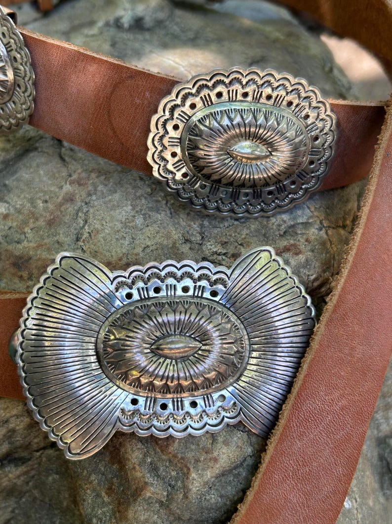 May include: Close-up of a brown leather belt with three silver-colored oval belt buckles. The buckles have intricate engraved designs with a central eye-shaped element and sunburst patterns. The belt is resting on a textured stone surface.