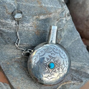 Puede incluir: Una petaca de plata con un dise&ntilde;o grabado y una piedra turquesa. La petaca tiene un peque&ntilde;o tap&oacute;n sujeto por una cadena. La petaca es redonda con un peque&ntilde;o pico. La petaca es de plata.