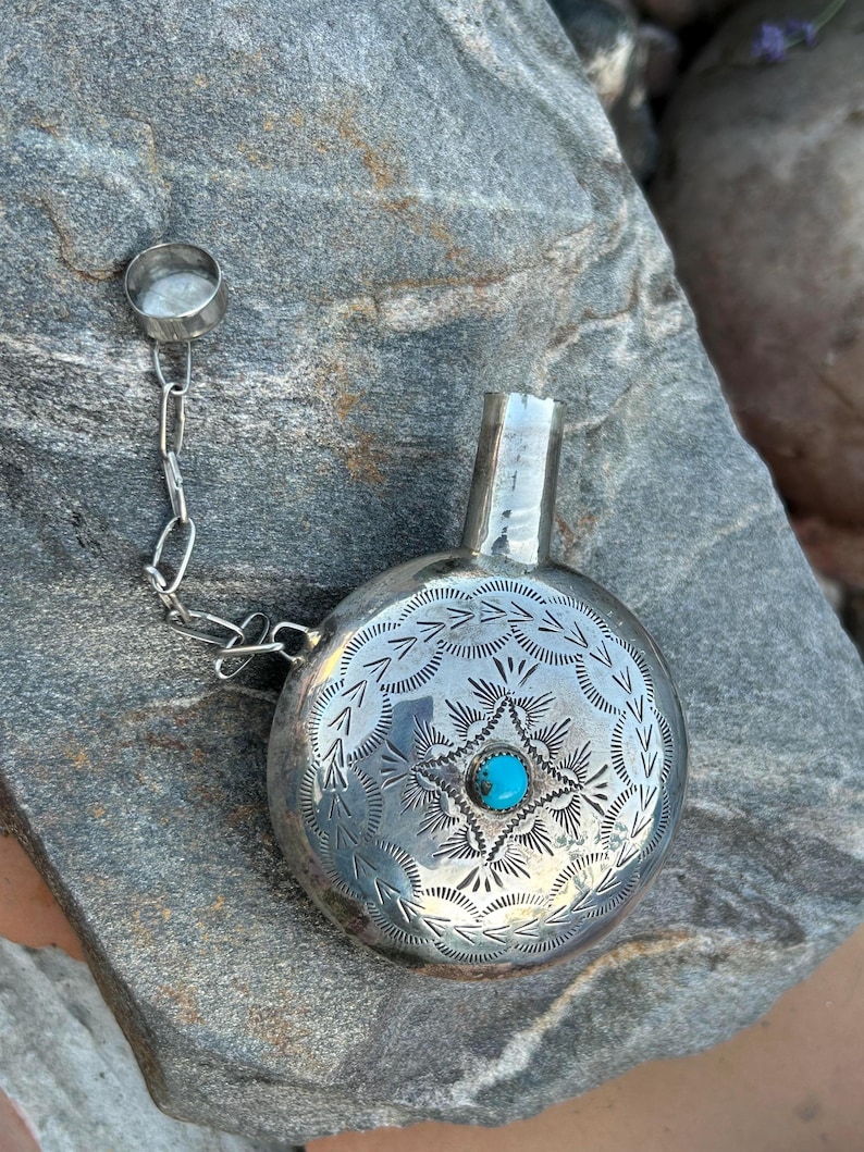 Puede incluir: Una petaca de plata con un dise&ntilde;o grabado y una piedra turquesa. La petaca tiene un peque&ntilde;o tap&oacute;n sujeto por una cadena. La petaca es redonda con un peque&ntilde;o pico. La petaca es de plata.