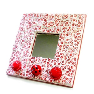 Puede incluir: Un marco de espejo cuadrado blanco con un diseño de mosaico floral rojo y blanco. Tres botones rojos con detalles negros están unidos al marco.