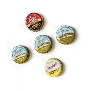 Könnte beinhalten: Fünf verschiedene Flaschenverschlüsse der Leinenkugel's Brauerei. Die Verschlüsse sind gelb, blau, rot und goldfarben mit verschiedenen Designs und Texten. Der Text auf den Verschlüssen umfasst "Leinie's", "Red Lager", "Summer Shandy" und "Helles Yeah".