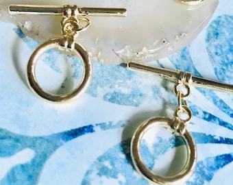 May include: Four gold-toned toggle clasps with round rings and a bar. The clasps are on a white agate surface.