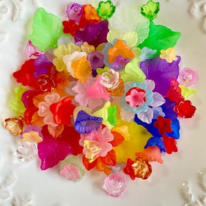 May include: A collection of colorful, translucent plastic flowers and leaves in various shapes and sizes. The assortment includes red, pink, yellow, orange, green, blue, and purple elements, creating a vibrant and cheerful display. The items are scattered on a white surface.