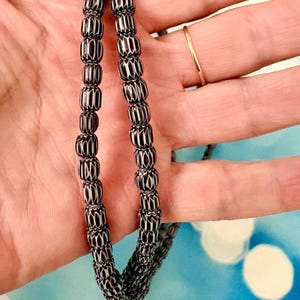 May include: A necklace composed of small, cylindrical black beads featuring a repeating white chevron or zigzag pattern. The beads are strung together in a continuous strand, displaying a consistent geometric design throughout the entire length of the piece.