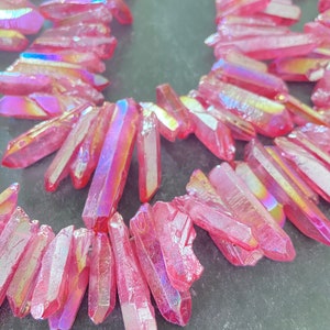 Può includere: Una fila di punte di cristallo rosa iridescente, note anche come quarzo aura angelica, disposte in un motivo decorativo. I cristalli sono sfaccettati e hanno un effetto scintillante, simile all'arcobaleno.