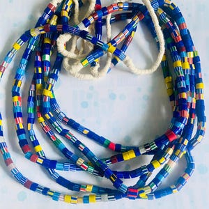 May include: A multi-strand necklace made of colorful glass beads. The beads are small and square-shaped, with a variety of colors including blue, yellow, red, and green. The necklace is strung on a white cord.