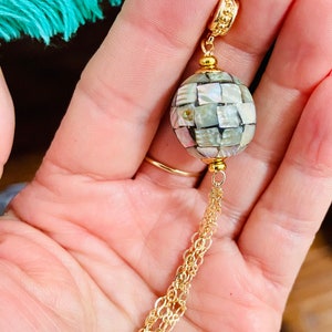 May include: A gold chain necklace with a large, round pendant made of white and gray mosaic tiles. The pendant is attached to the chain with a gold connector.