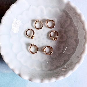 May include: Six small, circular gold-toned metal rings with a twisted rope-like texture. Each ring features a tiny, smooth spherical accent attached to the outer edge. These metallic components are arranged in a circular pattern on a white surface.
