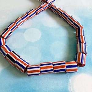 May include: A strand of cylindrical beads featuring a repeating striped pattern of vibrant orange, deep navy blue, and crisp white vertical lines. The beads are arranged in a loop, showcasing the consistent color sequence and smooth finish of each individual piece.