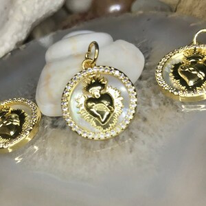 Peut inclure: Trois pendentifs dor&eacute;s avec un fond en nacre blanche. Chaque pendentif pr&eacute;sente une image d&eacute;taill&eacute;e d'un c&oelig;ur avec une couronne et une aur&eacute;ole. Les pendentifs sont entour&eacute;s d'un cercle de strass transparents.
