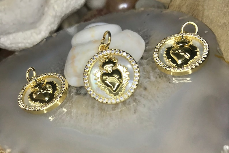 Peut inclure: Trois pendentifs dor&eacute;s avec un fond en nacre blanche. Chaque pendentif pr&eacute;sente une image d&eacute;taill&eacute;e d'un c&oelig;ur avec une couronne et une aur&eacute;ole. Les pendentifs sont entour&eacute;s d'un cercle de strass transparents.