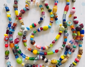 May include: A collection of multi-colored glass beads strung on clear nylon thread. The beads vary in shape, size, and pattern, featuring translucent, opaque, and striped designs in shades of blue, yellow, red, green, and purple, arranged in loose loops.