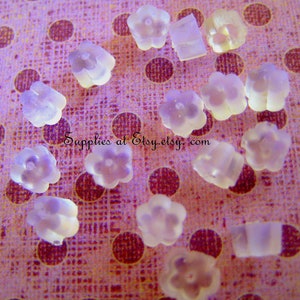 May include: A collection of translucent, flower-shaped earring backs and rectangular earring stoppers. The earring backs are scattered on a pink fabric with maroon polka dots. The text "Supplies at Etsy.etsy.com" is visible.