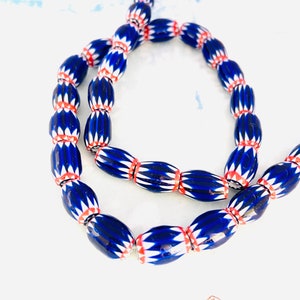 May include: A strand of blue, white, and red glass beads. The beads are oval-shaped with a dark blue base, white chevron pattern, and red accents. The beads are strung together, creating a necklace or bracelet.