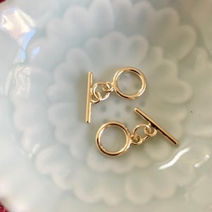 May include: Two gold-toned metal toggle clasps with a round ring and a bar. The clasps are on a white ceramic surface.