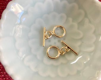 May include: Two gold-toned metal toggle clasps with a round ring and a bar. The clasps are on a white ceramic surface.