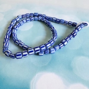 Pode incluir: Um fio de contas cilíndricas de vidro com um padrão repetitivo de riscas verticais azul-escuras e brancas. As contas estão enfiadas num laço contínuo, apresentando um design às riscas consistente em cada unidade de conta individual.