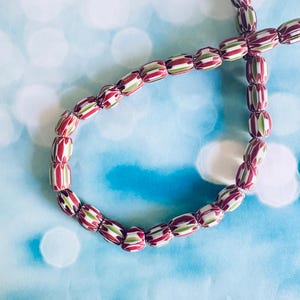 May include: Strung beads with a repeating pattern of red, green, and white stripes. The beads are oval-shaped and arranged in a curved line against a light blue background with white bokeh.