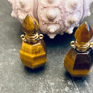 May include: Two small perfume bottles crafted from brown and gold tiger's eye gemstone. Each bottle features a faceted hexagonal body and a pointed gemstone stopper. The neck of each bottle is accented with a gold-toned metal collar and two small loops.