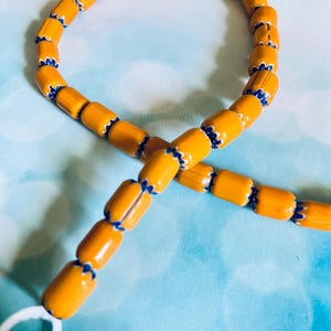 May include: A strand of orange beads with blue and white accents. The beads are cylindrical and strung together, forming a loop. The beads are on a light blue background.