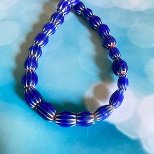 Puede incluir: Un collar de cuentas de vidrio azul con rayas blancas. Las cuentas ovaladas están ensartadas en un hilo rojo. El collar se muestra sobre un fondo azul claro.