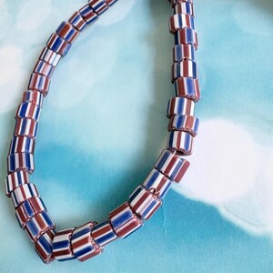 May include: A strand of cylindrical glass beads arranged in a repeating pattern of vertical stripes. Each bead features alternating bands of deep maroon, bright white, and royal blue, creating a consistent multi-coloured design throughout the strand.
