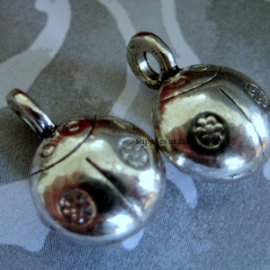 May include: Two silver-colored ladybug pendants with a loop for hanging. Each pendant has a rounded shape with etched details, including a line across the back and small circular designs. The pendants are on a patterned background.