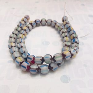 May include: A strand of round, white glass beads featuring a repeating pattern of brown star-shaped accents and thin, vertical stripes in shades of light blue, yellow, and green. The beads are strung together on a thin, light-colored thread.