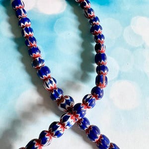 May include: A strand of blue, red, and white glass beads. The beads are oval-shaped with a chevron pattern in red and white against a blue background. The beads are strung together on a white string.
