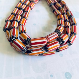 Può includere: Una collana a più fili con perline a righe colorate nei toni del rosso, arancione e bianco. Le perline cilindriche hanno dettagli neri alle estremità. La collana è presentata su una superficie chiara.