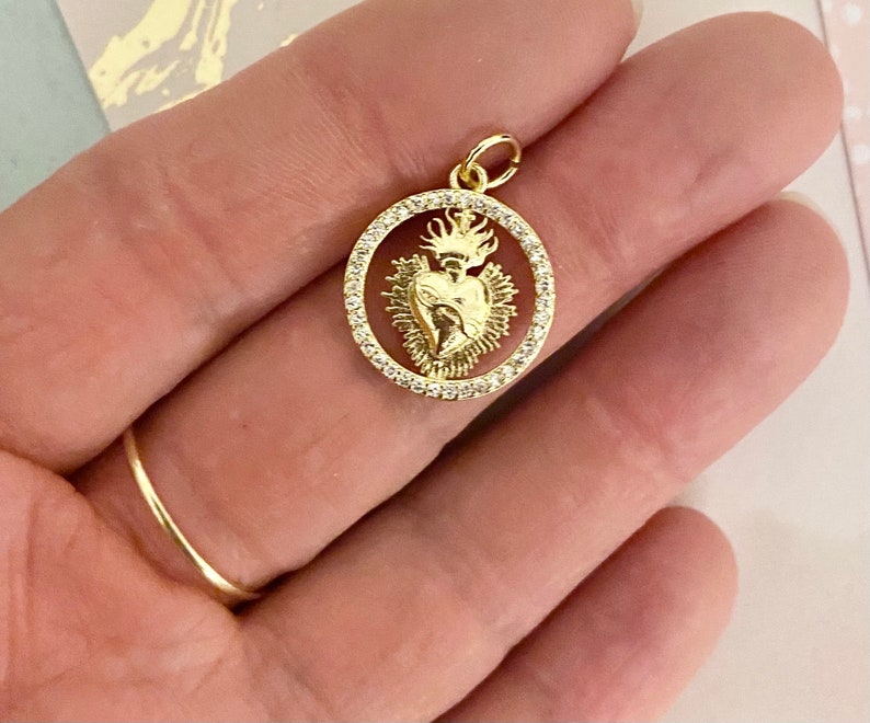 Peut inclure: Pendentif dor&eacute; repr&eacute;sentant une image d&eacute;taill&eacute;e d'un c&oelig;ur avec une couronne et des flammes, entour&eacute; d'un cercle de pierres brillantes transparentes.