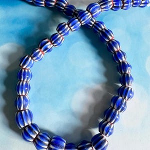 May include: A strand of blue and white striped glass beads. The beads are oval-shaped with a ribbed texture and are strung together with a thin red string. The beads are a deep blue color with white stripes and are arranged in a curved pattern.