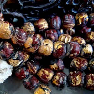 May include: Close-up of a collection of antique beads. The beads are a mix of burgundy, gold, and black. Some beads have a textured surface, and others have a smooth finish. The beads are strung together, creating a unique pattern.