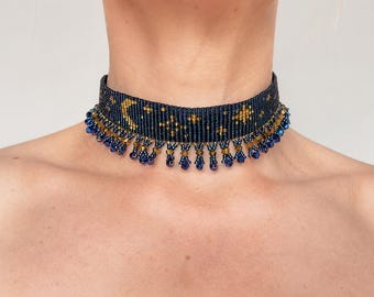 May include: A dark blue beaded choker necklace featuring a woven pattern of gold crescent moons and stars. The lower edge of the choker is decorated with a fringe of dangling dark blue and gold beads that hang in a repeating pattern around the neck.