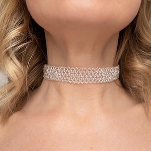 Könnte beinhalten: Nahaufnahme einer silbernen Choker-Halskette. Die Halskette besteht aus kleinen, miteinander verbundenen Perlen, die ein zartes, strukturiertes Aussehen erzeugen. Der Choker liegt um den Hals und zeigt sein kompliziertes Design und seine funkelnde Qualität. Der Hintergrund ist ein weicher, neutraler Ton.