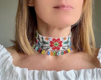 Może przedstawiać: Szeroki naszyjnik typu choker wykonany z białych koralików z kolorowym tkanym wzorem przedstawiającym czerwone kwiaty i zielone ptaki. Dolna krawędź naszyjnika wykończona jest frędzlami z wiszących, wielokolorowych koralików w kształcie łez.