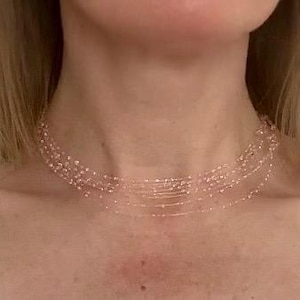 May include: A delicate, multi-strand necklace with small, light pink beads. The necklace is worn around the neck, creating a layered effect. The strands appear to be suspended, giving an airy, elegant look. The necklace is a jewelry item.