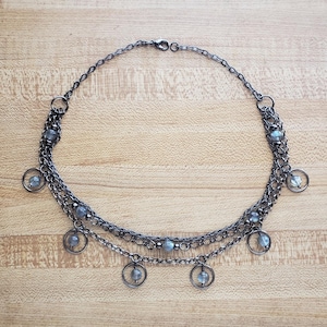 May include: A layered necklace with a gunmetal finish. The necklace features multiple strands of chain, adorned with small, round, light blue beads and dangling circular charms. The necklace is displayed on a light brown wooden surface.