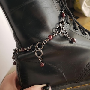 May include: A dark metal chainmail boot accessory featuring interconnected circular rings and small, round, deep red beads. The decorative piece is draped across the side of a smooth, black leather boot with black laces and yellow stitching.