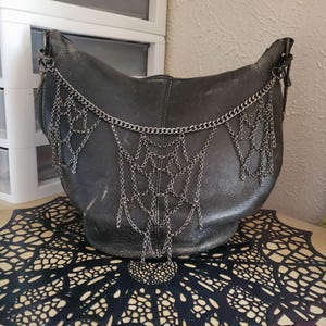 May include: A dark gray leather hobo bag featuring a decorative silver-toned metal chain arrangement draped across the front. The chains are linked together to form a web-like pattern that hangs from the top sides of the bag down to the center.