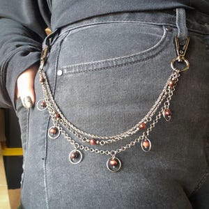 May include: Silver-toned chain with three strands, adorned with small brown beads and circular charms. The chain is attached to a pair of gray denim jeans with a silver clasp. A stylish accessory.