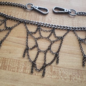 May include: A gunmetal-colored chain belt with a decorative, draped chain design. The belt features a series of small, dangling chains and two clasp closures. The belt is displayed on a light brown wooden surface.