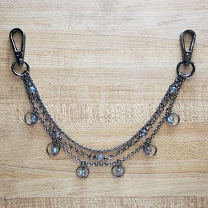 May include: Gunmetal-toned chain accessory with two carabiner clasps. The chain features three strands with small, round, light blue beads and dangling circular charms. The accessory is displayed on a light brown wooden surface.