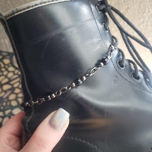 May include: A black leather boot with a silver chain ankle bracelet featuring black beads. The boot has black laces and a scuffed surface. The image is a close-up of the boot and ankle bracelet.