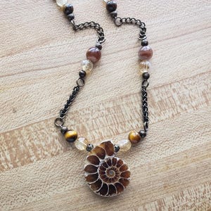 May include: A necklace featuring a central brown ammonite fossil pendant suspended from a dark metal chain. The chain is accented with alternating round beads in shades of brown, gold, and clear, including tiger's eye and quartz-like stones.