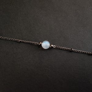 May include: A delicate dark metal chain necklace featuring a single round, translucent, pale blue moonstone bead at the center, flanked by two smaller, dark metallic spherical beads on either side of the central stone.