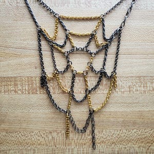 May include: A layered necklace with a web-like design. The necklace features alternating chains of gold and dark gray metal. The chains are connected to create a cascading effect, with some chains hanging freely. The necklace is displayed on a wooden surface.