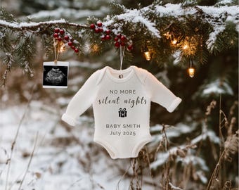 May include: A white baby onesie hanging from a snowy pine branch with Christmas lights. The onesie has the text "No more silent nights" and "Baby Smith July 2025" printed on it.